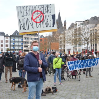 Ein junger Mann mit Mund-Nasen-Schutz hält ein selbst gemaltes Pappschild. Im Hintergrund viele weitere Demonstrant*innen und die Türme von Groß Sankt Martin in Köln.