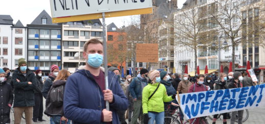 Ein junger Mann mit Mund-Nasen-Schutz hält ein selbst gemaltes Pappschild. Im Hintergrund viele weitere Demonstrant*innen und die Türme von Groß Sankt Martin in Köln.