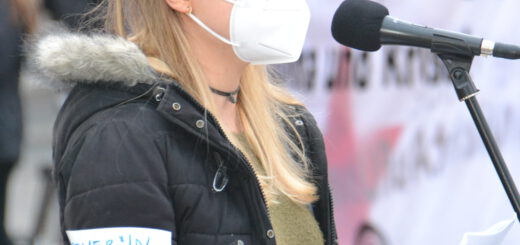 Eine junge Frau mit langen blonden Haaren, Brille und Mund-Nasen-Schutz spricht in ein Mikro. Sie trägt eine offene dunkle Winterjacke mit dünnem Fellkragen und hat eine Armbinde "Ordner*in"