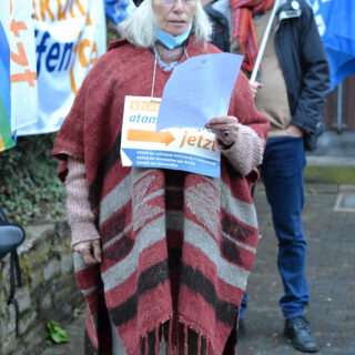 Eine ältere Dame mit dicken schulterlangen silbergrauen Haaren und schwarzer Tuchmütze in rötlichem Poncho liest von einem Blatt Papier. Im Hintergrund Transparente "Atomwaffenfrei jetzt".