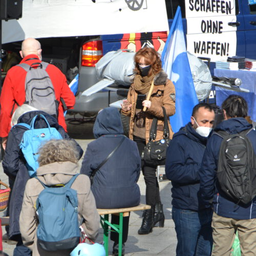Menschen in Winterkleidung wuseln vor einer improvisierten Bühne. Einige sitzen auf einer Bierbank. Alle tragen Mund-Nase-Masken. In der Mitte eine hübsche Frau, die lässig eine blaue Flagge trägt. Im Hintergrund eine Autoaufschrift "Frieden schaffen ohne Waffen".