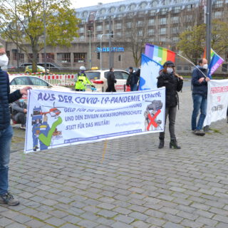 Fünf Leute mit Mund-Nasen-Schutz halten zwei Transparente und bunte Friedensfahnen am Rand eines gepflasterten Platzes.