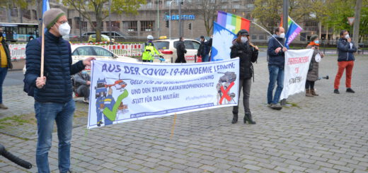 Fünf Leute mit Mund-Nasen-Schutz halten zwei Transparente und bunte Friedensfahnen am Rand eines gepflasterten Platzes.