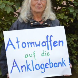 Eine hübsche ältere Dame mit schulterlangen silbergrauen Haaren und frischer Gesichtsfarbe hält fröhlich schmunzelnd ein selbst gemaltes Plakat vor sich: "Atomwaffen auf die Anklagebank". Im Hintergrund eine sommergrüne Buchenhecke.