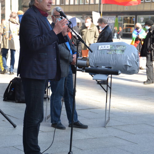 Ein großer Mann mit grauen Haaren und dunklem Blazer spricht in ein Mikro. Im Hintergrund das Modell einer Kampfdrohnen und zuhörende Menschen.