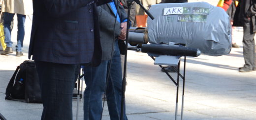 Ein großer Mann mit grauen Haaren und dunklem Blazer spricht in ein Mikro. Im Hintergrund das Modell einer Kampfdrohnen und zuhörende Menschen.