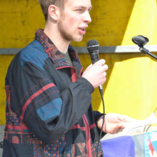 Ein junger Mann mit hellem Bart in bunter Windjacke spricht in ein Mikro.