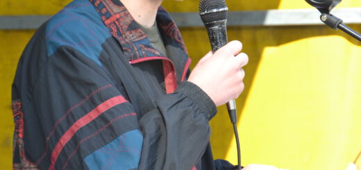 Ein junger Mann mit hellem Bart in bunter Windjacke spricht in ein Mikro.