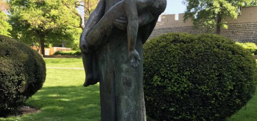 Bronzeskulptur einer Frau, die einen toten Jungen im Arm trägt. Im Hintergrund sind Parkbäume und ein Teil der Kölner Stadtmauer zusehen.