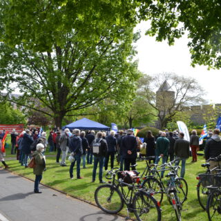 Menschen mit Fahnen gruppieren sich um einen Mittelpunkt in einem sonnigen grünen Park. Im Vordergrund auf einer Wiese abgestellte Fahrräder.