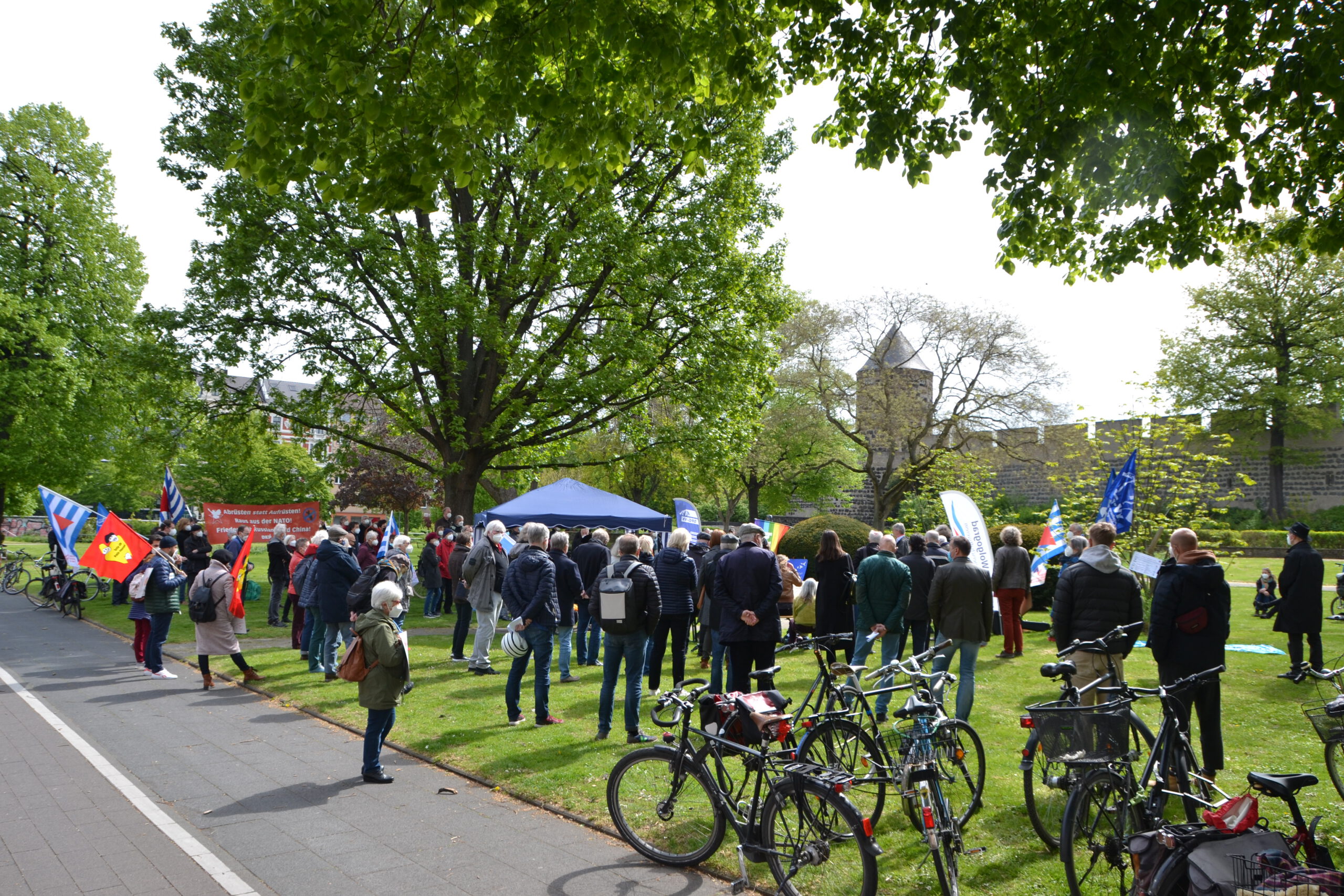 Menschen mit Fahnen gruppieren sich um einen Mittelpunkt in einem sonnigen grünen Park. Im Vordergrund auf einer Wiese abgestellte Fahrräder.