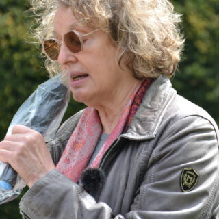 Eine Frau mit Sonnenbrille und dichten grau-blonden kinnlangen Locken spricht in ein Mikrofon, das in eine Plastikfolie eingewickelt ist. Sie trägt ein rosa Tuch und eine erdgraue Lederjacke. Im Hintergrund grünes Blattwerk.