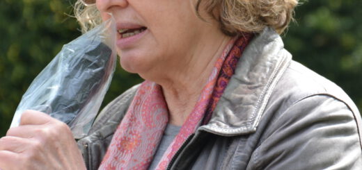 Eine Frau mit Sonnenbrille und dichten grau-blonden kinnlangen Locken spricht in ein Mikrofon, das in eine Plastikfolie eingewickelt ist. Sie trägt ein rosa Tuch und eine erdgraue Lederjacke. Im Hintergrund grünes Blattwerk.