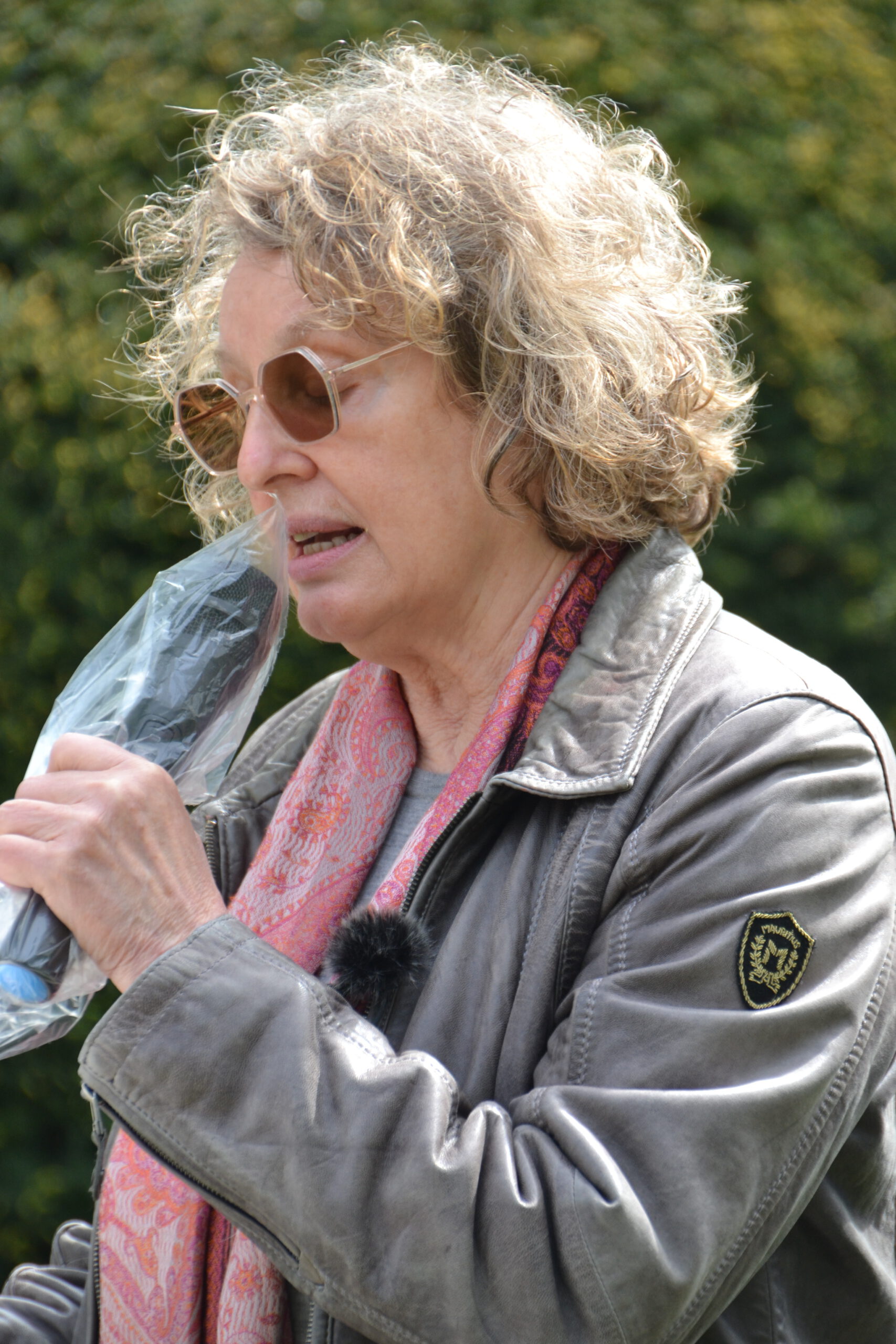 Eine Frau mit Sonnenbrille und dichten grau-blonden kinnlangen Locken spricht in ein Mikrofon, das in eine Plastikfolie eingewickelt ist. Sie trägt ein rosa Tuch und eine erdgraue Lederjacke. Im Hintergrund grünes Blattwerk.