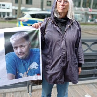 Eine grauhaarige Dame hält das Fotoporträt eines kräftigen Mannes mit graumelierten kurzen Haaren in einem blauen T-Shirt.