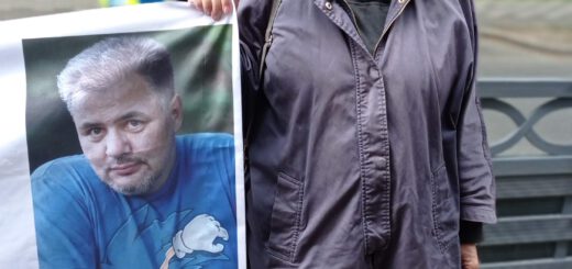 Eine grauhaarige Dame hält das Fotoporträt eines kräftigen Mannes mit graumelierten kurzen Haaren in einem blauen T-Shirt.