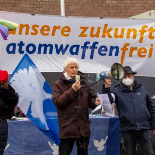 Ein schlanker weißhaariger Mann in braunem Winteranorak spricht in ein Megafon. Im Hintergrund ein weißes Transparent mit orangefarbener und dunkelblauer Schrift "unsere zukunft atomwaffenfrei". Neben ihm zwei weitere Personen, sowie weiße Friedenstauben auf blauen Flaggen.