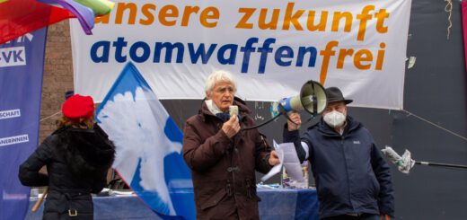 Ein schlanker weißhaariger Mann in braunem Winteranorak spricht in ein Megafon. Im Hintergrund ein weißes Transparent mit orangefarbener und dunkelblauer Schrift "unsere zukunft atomwaffenfrei". Neben ihm zwei weitere Personen, sowie weiße Friedenstauben auf blauen Flaggen.