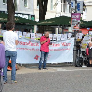 Ein Mann in pinkem Hemd und Jeans spricht vor einem weißen Transparent mit der Aufschrift "Gesundheit ist keine Ware". Umstehende klatschen.