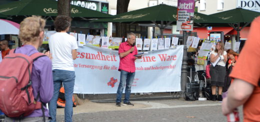 Ein Mann in pinkem Hemd und Jeans spricht vor einem weißen Transparent mit der Aufschrift "Gesundheit ist keine Ware". Umstehende klatschen.