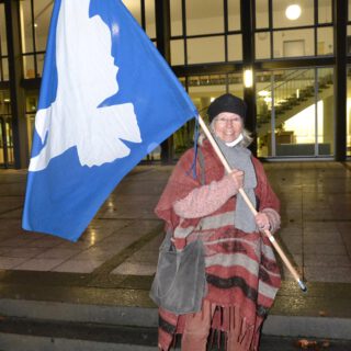Eine ältere Dame in rotbrauner Hose und rotbraun-grau-gestreiftem Poncho mit schwarzer Mütze hält eine große Flagge mit weißer Friedenstaube auf blauem Grund und strahlt in die Kamera. Es ist dunkel. Hinter ihr ein von innen hell erleuchtetes Verwaltungsgebäude, in dem eine Halle mit Treppenaufgängen zu erkennen ist.