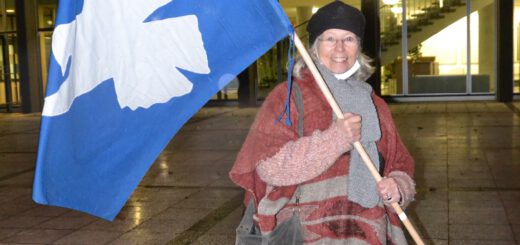 Eine ältere Dame in rotbrauner Hose und rotbraun-grau-gestreiftem Poncho mit schwarzer Mütze hält eine große Flagge mit weißer Friedenstaube auf blauem Grund und strahlt in die Kamera. Es ist dunkel. Hinter ihr ein von innen hell erleuchtetes Verwaltungsgebäude, in dem eine Halle mit Treppenaufgängen zu erkennen ist.