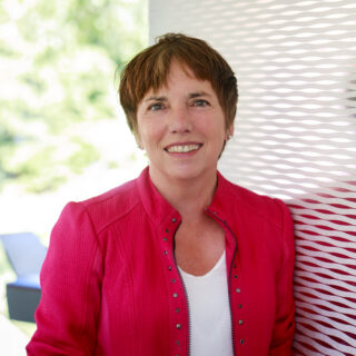 Eine Dame mit kurzen dunklen Haaren in roter sportlich-schicker Jacke und weißem Shirt schaut freundlich in die Kamera.