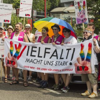 Eine Gruppe bunt und sommerlich gekleideter Demonanstrant*innen zeigen fröhlich Schilder und ein Transparent "Vielfalt macht uns stark! LSVD NRW - Die Lobby für Lesben & Schwule"