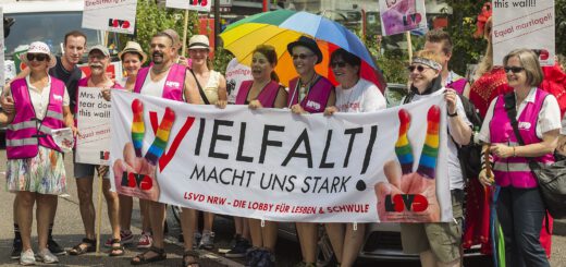 Eine Gruppe bunt und sommerlich gekleideter Demonanstrant*innen zeigen fröhlich Schilder und ein Transparent "Vielfalt macht uns stark! LSVD NRW - Die Lobby für Lesben & Schwule"