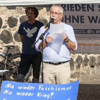 Ein älterer Herr in heller Sommerhose und hellblauem Hemd spricht in ein Mikrofon. Im Hintergrund ein junger Mann mit blauem T-Shirt, auf dem eine weiße Friedenstaube zu sehen ist. Vorn ist ein blaues selbst beschriftetes Plakat mit der Aufschrift "Nie wieder Faschismus! Nie wieder Krieg!" zu sehen.
