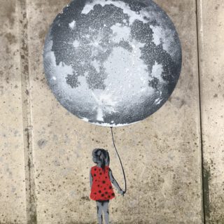 Ein kleines Mädchin in rotem Sommerkleid schaut verträumt vom Betrachter weg, während es eine große grau-weiße Weltkugel wie einen Luftballon an einem Faden hält. Bild auf dem Fuß einer Betonwand angebracht.