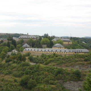 Blick über waldige Mittelgebirgshöhen. Die Anhöhe im Vordergrund ist mit größeren Gebäuden mit Satteldächern und Dachgauben bebaut.