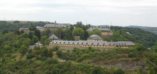 Blick über waldige Mittelgebirgshöhen. Die Anhöhe im Vordergrund ist mit größeren Gebäuden mit Satteldächern und Dachgauben bebaut.