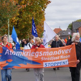 Zwei Frauen und ein Mann tragen ein orange-blau-weißes Transparent. Dahinter ein Demozug mit Friedensfahnen vor einer Reihe Backsteinhäusern und herbstlichen Bäumen.