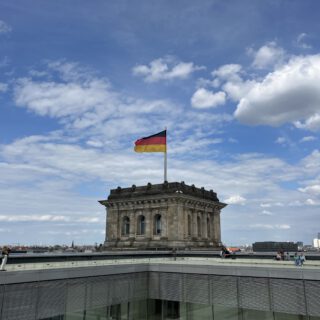 Im Zentrum das obere Ende eines massiven Vierkantturms im Stil der Neorenaissance mit Deutschland-Flagge vor einem blauen Himmel mit weißen Wolken. Im Hintergrund ist ein schlanker Fernsehturm mit einer glänzenden Kugel zu sehen (ehem. Fernsehturm der DDR auf dem Alexanderplatz).