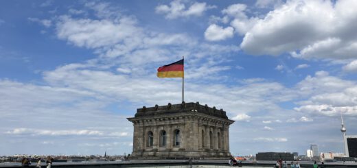 Im Zentrum das obere Ende eines massiven Vierkantturms im Stil der Neorenaissance mit Deutschland-Flagge vor einem blauen Himmel mit weißen Wolken. Im Hintergrund ist ein schlanker Fernsehturm mit einer glänzenden Kugel zu sehen (ehem. Fernsehturm der DDR auf dem Alexanderplatz).