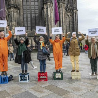 Acht Leute, zum Teil in orangefarbenen Overalls, stehen nebeneinander auf je einem leeren Getränkekasten und halten je ein Schild in die Höhe mit den Aufschriften "Wissen", "Respekt", "Freiheit", "Julian Assnage", "Kunstfreiheit", "Menwschenrechte", "Frieden". Im Hintergrund ist das Hauptportal des Kölner Doms zu sehen.