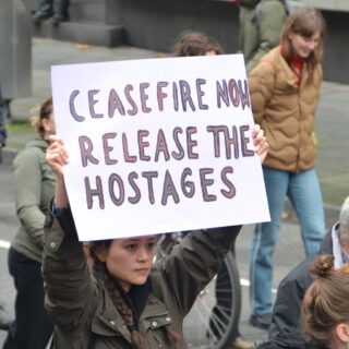 Eine ernste junge Frau in brauner Jacke hält in einer Gruppe laufender Menschen ein selbstgemaltes Schild in die Höhe.