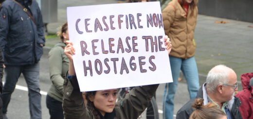 Eine ernste junge Frau in brauner Jacke hält in einer Gruppe laufender Menschen ein selbstgemaltes Schild in die Höhe.