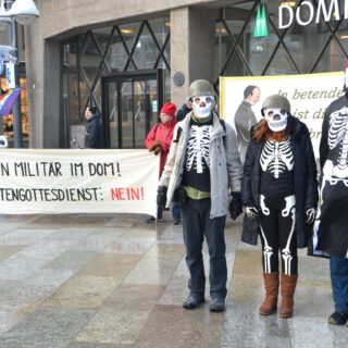 Drei als Skelette mit Soldatenhelm verkleidete Menschen vor einem Transparent.