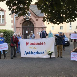 Ein Dutzend meist älterer Männer und Frauen, zum Teil mit Mund-Nase-Schutzmasken, stehen mit einem selbstgemalten weißen Transparent "Atombomben auf die Anklagebank!" vor einem schönen alten Gebäude aus rotem Sandstein.