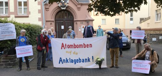 Ein Dutzend meist älterer Männer und Frauen, zum Teil mit Mund-Nase-Schutzmasken, stehen mit einem selbstgemalten weißen Transparent "Atombomben auf die Anklagebank!" vor einem schönen alten Gebäude aus rotem Sandstein.