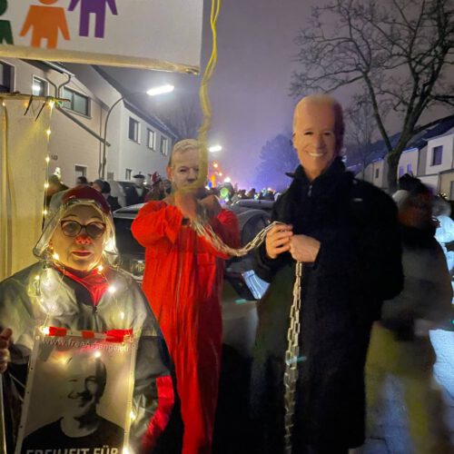 Als Joe Biden verkleidete Person hält eine Kette, mit der "Julian Assange" in orangefarbener Gefängnis-Kluft gefesselt ist. Im Vordergrund eine Frau mit Clown-Nase und Lichterkette vor einem nebligen Abendhimmel und anderen Leuten.