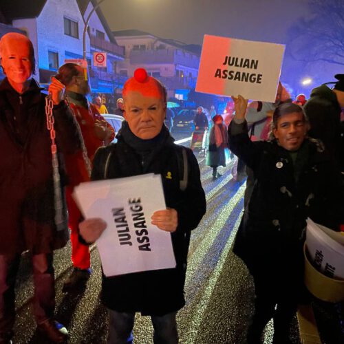 Joe Biden mit Olaf Scholz und Rishi Sunak, die je ein "Julian Assange"-Schild halten. Im Hintergrund eine nächtliche, neblige Straße mit weiteren Leuten.