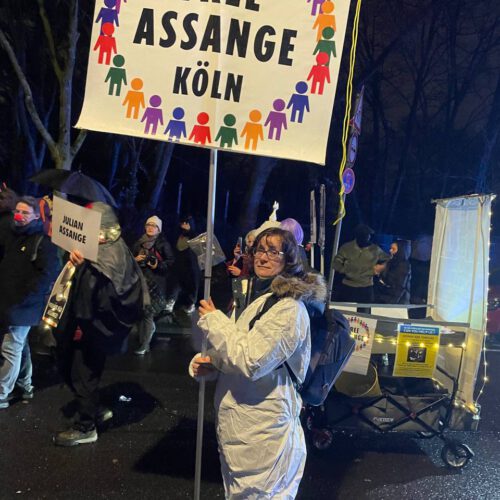 Eine Frau in weißem Overall hält ein großes Schild "Free Assange Köln".
