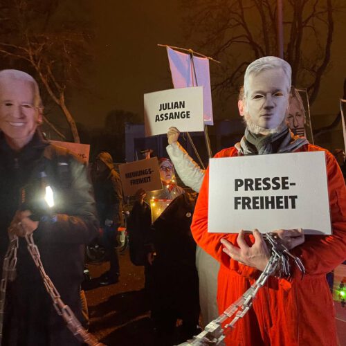 Hinter Joe Biden und Julian Assange sind weitere Leute zu sehen, die "Julian-Assange"-Schilder hochhalten