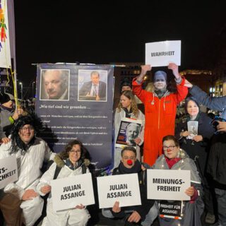 Ein knappes Dutzend gut gelaunte Aktivist*innen, als Julian Assange, Joe Biden, Annalena Baerbock, Gefangene und Clowns verkleidet, zeigen ihre Schilder und Plakate.