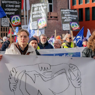 Ein junger Mann und eine junge Frau tragen ein helles Transparent mit der künstlerischen Darstellung einer Friedenstaube und verschränkten Händen. Dahinter viele Leute mit Demoschildern.