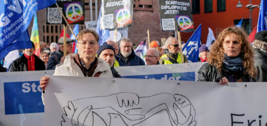 Ein junger Mann und eine junge Frau tragen ein helles Transparent mit der künstlerischen Darstellung einer Friedenstaube und verschränkten Händen. Dahinter viele Leute mit Demoschildern.