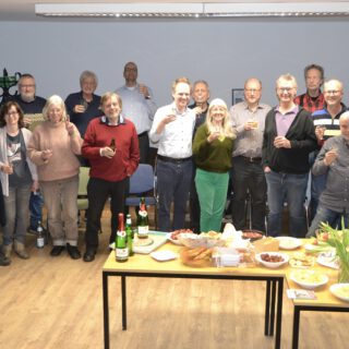 Anderthalb Dutzend Erwachsene stehen gut gelaunt mit Sektgläsern hinter einem gedeckten Tisch.